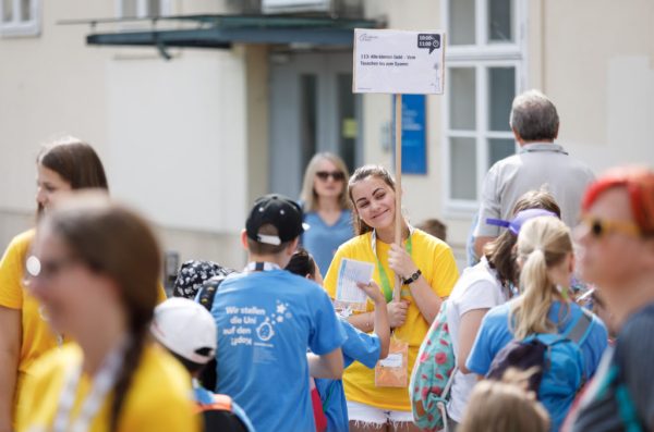 ein Teammitglied an der KinderuniWien steht in der Menschenmenge und hält lächelnd ein Lehrveranstaltungsschild hoch