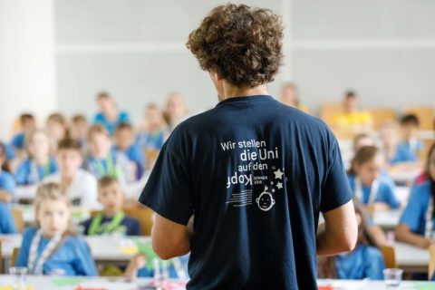 Ein KinderuniWien-Lehrender in seiner Lehrveranstaltung, man sieht seinen Rücken mit der T-Shirt Aufschrift "Wir stellen die Uni auf den Kopf"