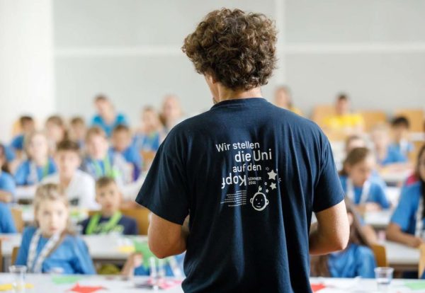 Ein KinderuniWien-Lehrender in seiner Lehrveranstaltung, man sieht seinen Rücken mit der T-Shirt Aufschrift "Wir stellen die Uni auf den Kopf"
